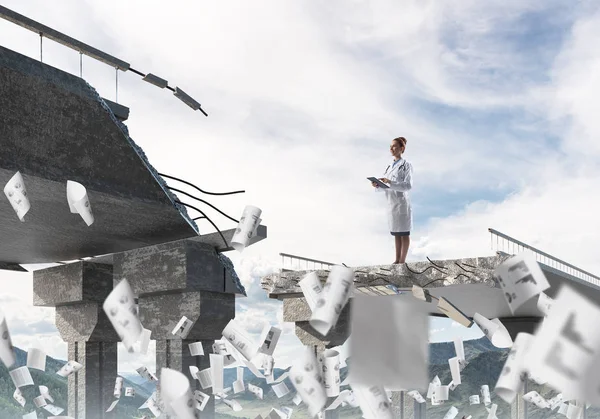 背景に風景ビューと壊れた橋の端に飛んで紙の中に立って白い医療の制服の若い女性医師 医療業界のコンセプト — ストック写真