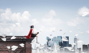 Maskeli ve pelerinli küçük bir çocuk çatıda oturmuş kitap okuyor. Karışık ortam