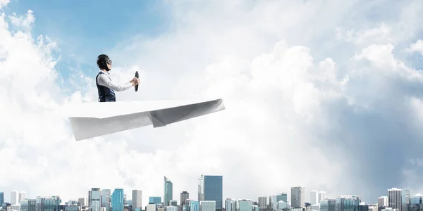 メガポリスの上の紙飛行機を運転する飛行士 紙飛行機に座り ハンドルを握っているパイロットの側面図 高層ビルやオフィスビルが立ち並む街並み ミックスメディアビジネスコンセプト — ストック写真