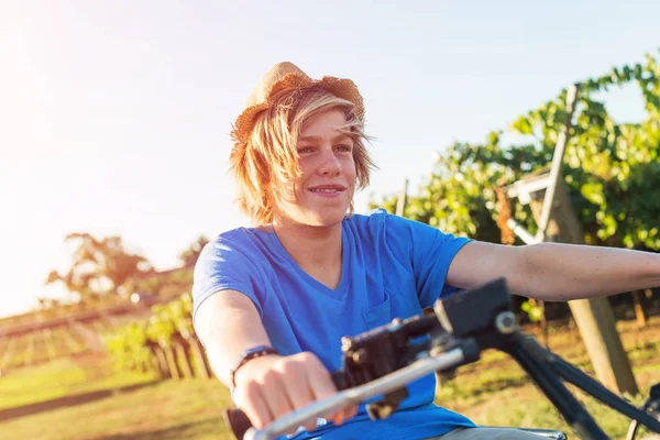 Young farmer riding quad bike on background of vine rows. Traditional farming in vineyard. Happy blond guy in straw hat and and blue t-shirt sitting on bike. Ecological food production in countryside.