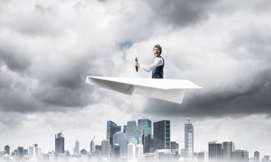 Havacı şapkalı işadamı ve gözlükler fırtınada kağıt uçak sürüyor. Kriz yönetimi kavramı. Küçük kağıt uçak pilot yan görünümü. Dramatik karanlık bulutlu gökyüzü ile Megalopolis panorama.