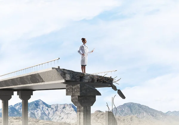 背景に風景ビューと壊れた橋の端に立っている白い無菌スーツの女性医師の概念画像 医療業界のコンセプト — ストック写真