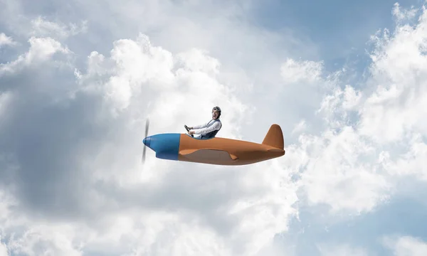 Man in aviator hat with goggles driving propeller plane. Funny man having fun in small airplane. Blue cloudy sky with fluffy clouds. Businessman sitting in paper plane and holding steering wheel.