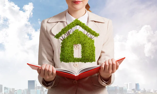Woman with green plant shaped house above opened notebook. Real estate agency advertising. Sale new real property. Elegant young woman in white business suit on background of cityscape and blue sky.