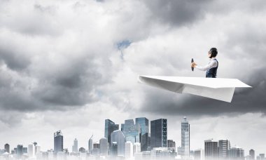 Havacı şapkalı işadamı ve gözlükler fırtınada kağıt uçak sürüyor. Kriz yönetimi kavramı. Küçük kağıt uçak pilot yan görünümü. Dramatik karanlık bulutlu gökyüzü ile Megalopolis panorama.