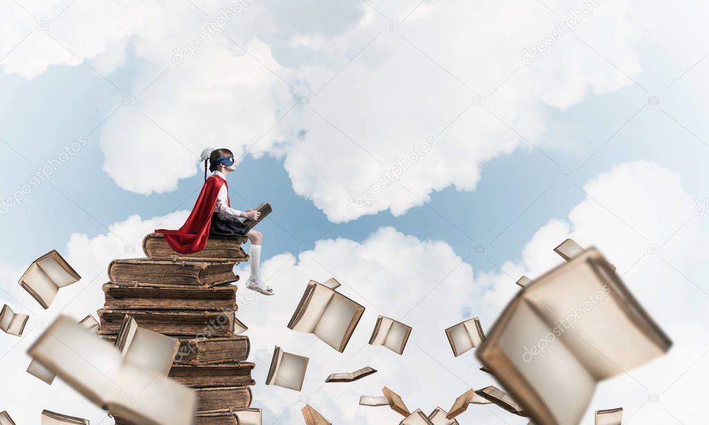 Little child in mask and cape sittting on pile of old books in sky and reading