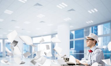 Genç yazar şapka ve gözlük kağıtları uçan arasında kapalı ve arka plan üzerinde office manzaralı masada otururken yazarak makinasıyla adam.