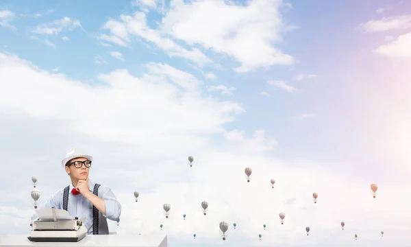 Young man writer in hat and eyeglasses using typing machine while sitting at the table with flying aerostats cloudy skyscape on background.
