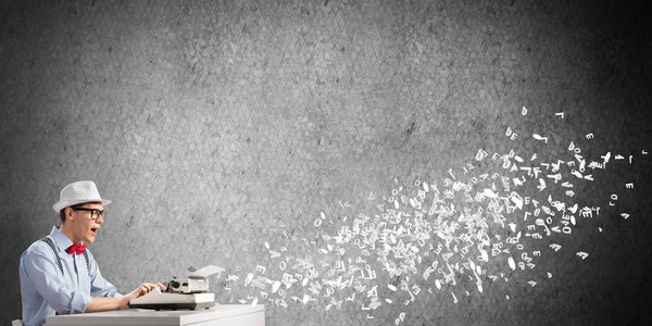 青年作家在帽子和眼镜使用打字机坐在桌之间在飞行信件和反对灰色混凝土墙壁在背景上 — 图库照片