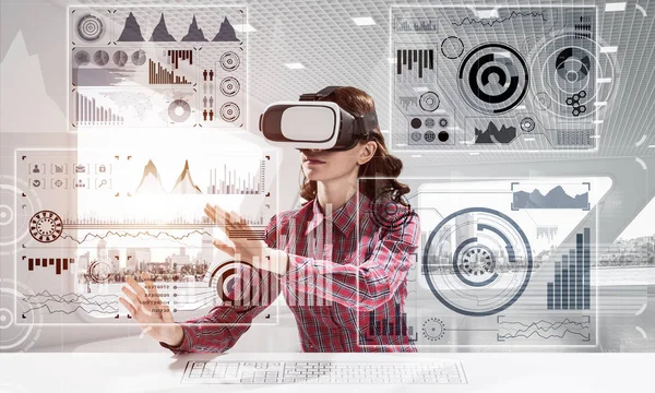 Educational process with help of VR headset. Cheerful young woman in checkered shirt using digital media interface while sitting indoors of office building.