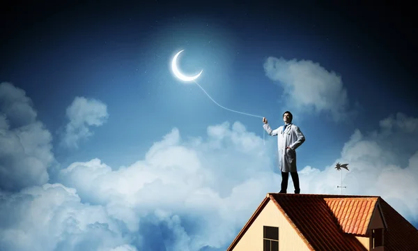 自信的医生站在砖屋顶顶部反对夜景和深蓝色天空的概念图 背景上有年轻的月亮 — 图库照片