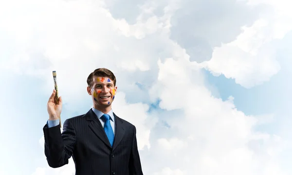 年轻而成功的商人穿着黑色西装 手里拿着画笔 微笑着 而站在背景上的多云的蓝色天空的概念形象 — 图库照片