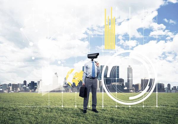 Camera headed man standing on green grass against modern citysca — Stock Photo, Image