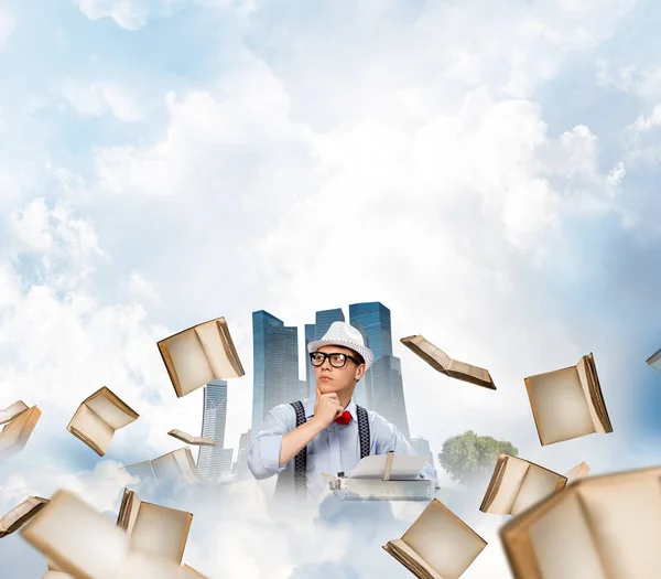 帽子と眼鏡の背景に水上都市と曇り空の景色の本を飛行中テーブルに座ってタイピング マシンを使用して若い男作家 — ストック写真