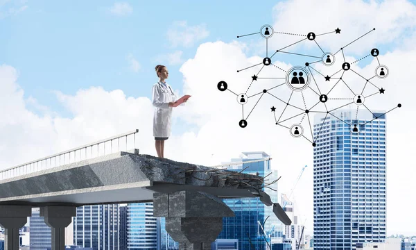 Confident female doctor in white medical suit looking at social network structure while standing at the end of broken bridge. Medical industry concept. Cityscape view on background