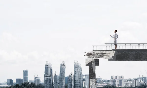 Confident medical industry employee in white medical suit holding tablet in hands while standing on broken bridge with cityscape view on background. Medical industry concept