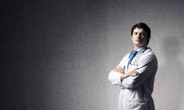 Portrait confident and professional man doctor in white medical suit keeping arms crossed and looking to camera while standing against dark gray wall on background. Medical industry employee concept