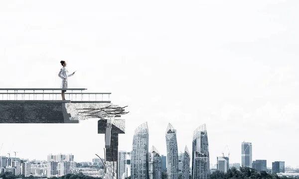 Confident medical industry employee in white medical suit holding tablet in hands while standing on broken bridge with cityscape view on background. Medical industry concept