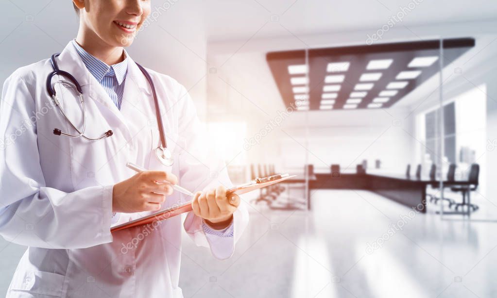 Side view of confident woman doctor in white sterile suit writing in notebook while standing indoors of white hospital building with sunlight on background. Medical industry concept