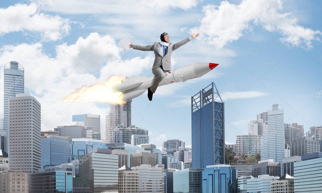 Conceptual image of young and happy businessman in suit flying on rocket with modern cityscape with skyscrapers and blue sky on background.