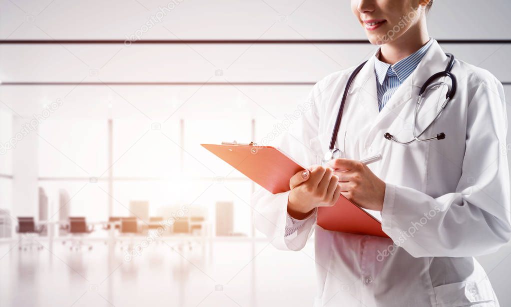 Close up of confident female doctor in white medical uniform making notes into notebook while standing inside of bright hospital building. Medical industry concept