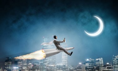 Arka plan üzerinde moon ile gece cityscape ve mavi karanlık gökyüzü ile roket üzerinde uçan takım elbiseli genç işadamı kavramsal görüntü.