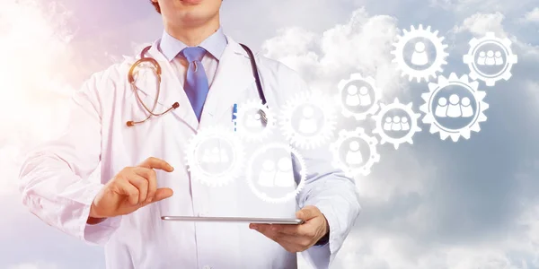 年轻医生穿着白色医疗服 手拿着带有团队齿轮结构的平板电脑的概念形象 同时站在背景上面对多云的风景景观 — 图库照片