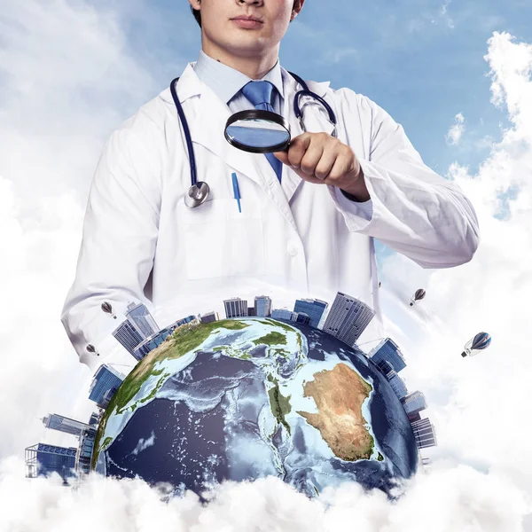 Cropped image of professional man doctor in white suit and stethoscope on neck discovering Earth globe through magnifier, with cloudy skyscape on background. Medical industry concept. Elements of this image furnished by NASA