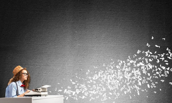 Young and beautiful woman writer in hat and eyeglasses using typing machine while sitting at the table among flying letters and against gray concrete wall on background.