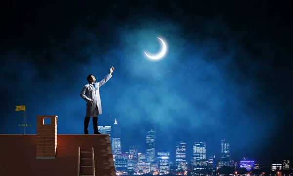 成功的医生在白色医疗制服看着年轻的月亮 而站在砖屋顶和反对夜景在背景上的看法 — 图库照片
