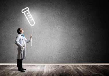 Arka planda koyu gri duvara karşı parlayan şişe sembolü whie ayakta beyaz Tıbbi üniforma genç güvenilir doktor yatay çekim.