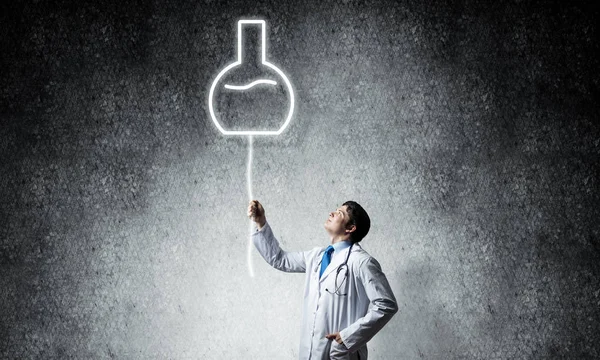年轻自信的医生在白色医疗制服插带发光的小瓶符号在背景的深灰色墙壁站立在 — 图库照片