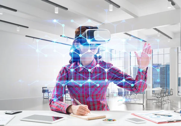 Parlak ofis binası içinde otururken sanal gerçeklik kulaklık ve medya arayüzü ile interracting kullanarak damalı gömlek genç ve güzel kadın kavramsal görüntü.