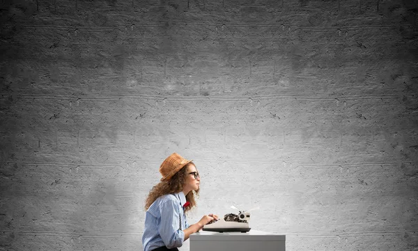 年轻美丽的女作家在帽子和眼镜的侧面观看使用打字机坐在桌子对面的灰色混凝土墙上的背景 — 图库照片