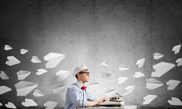 stock image Young man writer in hat and eyeglasses using typing machine while sitting at the table among flying paper planes and against gray concrete wall on background.