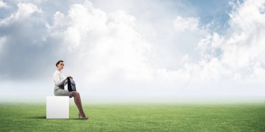 Elinde bavuluyla beyaz küpün üzerinde oturan zarif bir iş kadını.