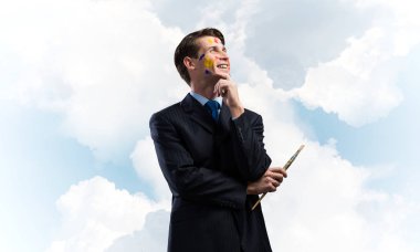 Boya fırçası elinde tutan ve uzak duran mavi bulutlu gökyüzü arka plan üzerinde karşı arayan genç düşünceli adam portresi.