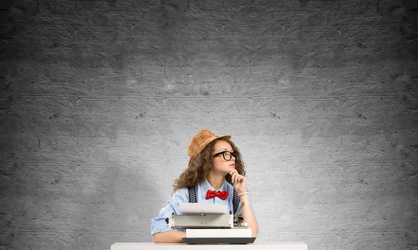 年轻美丽的女作家在帽子和眼镜使用打字机 而坐在桌子上反对灰色混凝土墙上的背景 — 图库照片