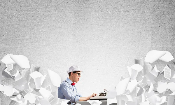 Young man writer in hat and eyeglasses using typing machine while sitting at the table among flying papers and against gray concrete wall on background.