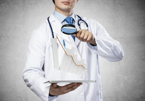 年轻的医生在无菌医疗服提出片剂与图形图表在手 而站在深灰色的墙壁在背景 — 图库照片
