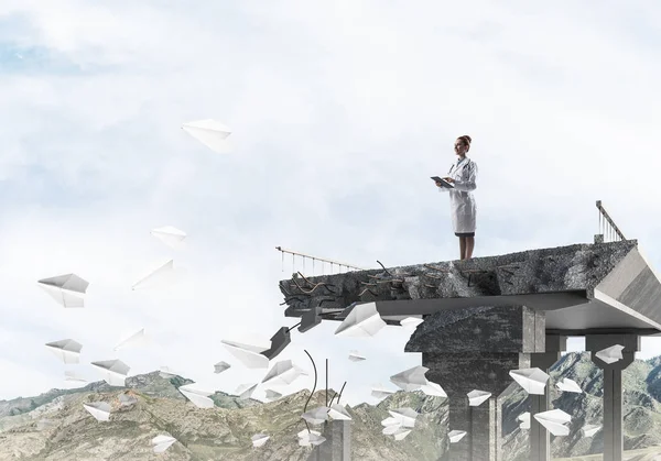 Professional medical industry employee using tablet while standing among flying paper planes at the edge of broken bridge with landscape view on background. Medical industry concept