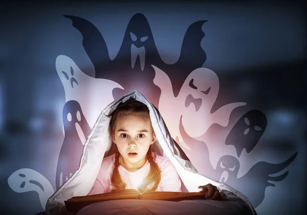 Scared kid with reading book under blanket. Afraid girl lying in bed at home. Child reading scary stories. Girl in pajamas and imaginary ghosts back on night sky. Bright light shining from open book.