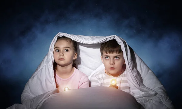 有手电筒的孩子躺在床上 小妹妹和弟弟一起躲在毯子下 在深蓝色的天空背景下 孩子们害怕夜晚的黑暗 儿童夜惊 — 图库照片