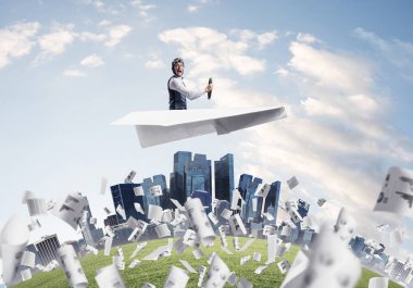 İş merkezinin üzerinde etkileyici havacı sürüş kağıt uçak. Deri kasklı pilot kağıt düzlemde oturuyor. Düşen kağıt kağıtlar. Yüksek gökdelenler ve ofis binaları ile şehir silueti yuvarlak.