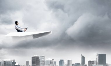 Havacı şapkalı işadamı ve gözlükler fırtınada kağıt uçak sürüyor. Kriz yönetimi kavramı. Küçük kağıt uçak pilot yan görünümü. Dramatik karanlık bulutlu gökyüzü ile Megalopolis panorama.