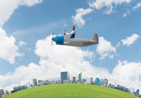 尖叫的商人在皮革头盔飞行在螺旋桨飞机 情绪飞行员驾驶小型飞机在城市景观之上 圆润的城市天际线与绿草 蓝天和现代摩天大楼 — 图库照片