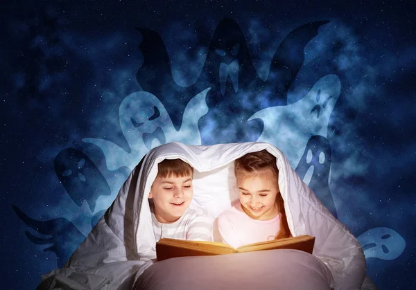 快乐的小孩在床上读魔法童话 哥哥和妹妹一起躲在毯子下 小朋友在睡衣与打开的大书和假想的怪物在深蓝色的背景 — 图库照片