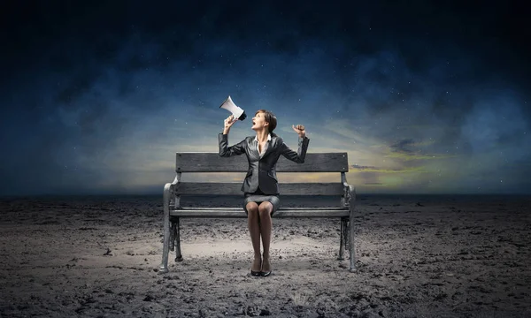 Business woman with megaphone sitting on wooden bench. Female speaker shouting in loudspeaker outdoors. Business marketing and promotion. Beautiful skyscape with deep blue night sky.