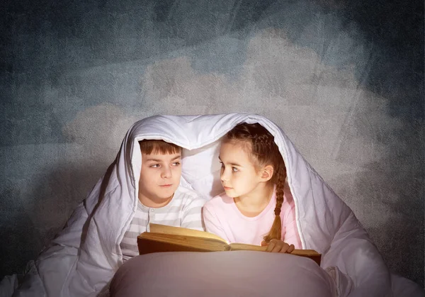 小さな女の子と男の子が毛布の下でお互いを見ている 寝る前にベッドで魔法の物語を読む子供たち 灰色の壁を背景にパジャマの妹と兄弟 — ストック写真