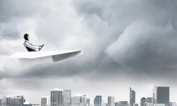 Businessman in aviator hat and goggles driving paper plane in storm. Crisis management concept. Side view of pilot in small paper airplane. Megalopolis panorama with dramatic dark cloudy skyscape.